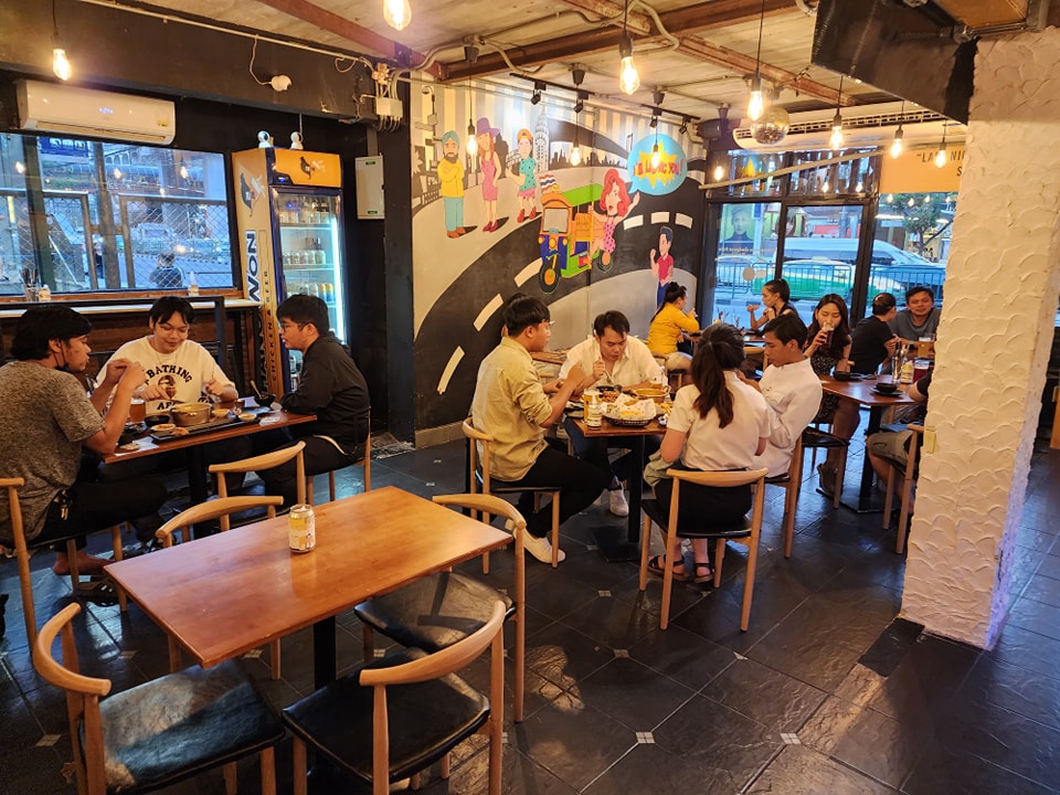 The lively interior of the restaurant with customers dining at wooden tables. The decor features industrial-style lighting and a large, colorful mural depicting a street scene with a Tuk-Tuk and various characters.